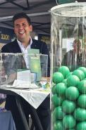Ein Mann lächelt hinter einem Messestand, neben einem großen Display mit mehreren leuchtend grünen Kugeln.