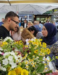Nahaufnahme von zahlreichen frischen gelben und weißen Blumen im Vordergrund, umgeben von einer Gruppe von Menschen bei einem Markt.