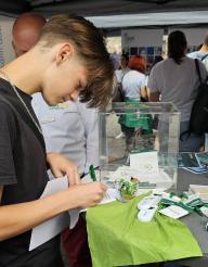 Ein junger Mann sitzt an einem Stand und bearbeitet Dokumente, während im Hintergrund weitere Personen bei einem Treffen interagieren.