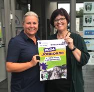 Zwei lächelnde Frauen stehen neben einem großen Werbeplakat für die Jobbörse 2024.