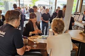 Mehrere Menschen stehen und sitzen in einem belebten Außenbereich, der wie ein Markt oder ein Café wirkt.