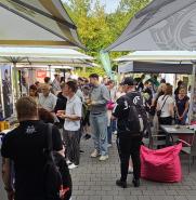Ein belebtes Außenmarkttreiben mit zahlreichen Menschen, die zwischen weißen und dunklen Verkaufszelten spazieren.
