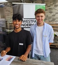 Zwei junge Männer stehen vor einem Banner mit dem Schriftzug Westfanden und lächeln in die Kamera.