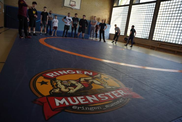 Innenaufnahme eines Wrestling-Trainingsbereichs mit mehreren Sportlern auf der markierten blauen Matte.