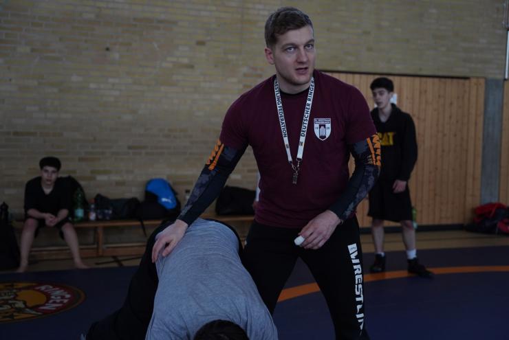 Ein junger Mann in Sportkleidung steht in einer Sporthalle, umgeben von anderen Sportlern und Trainingsmaterialien.