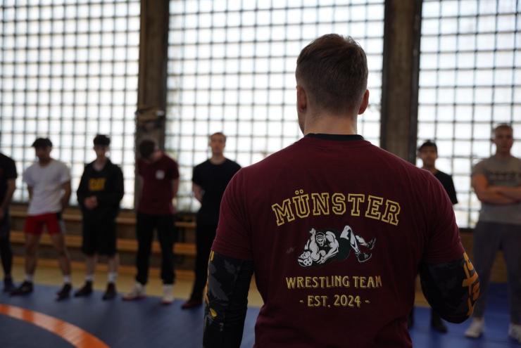 Ein Sportler steht im Vordergrund mit dem Rücken zur Kamera und trägt ein Trikot des Münster Wrestling Teams. Im Hintergrund sind weitere Personen in 