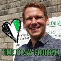 Ein lächelnder Mann steht vor einem Schild, auf dem die Worte TIME TO SAY GOODBYE! stehen.