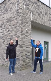 Zwei Männer posieren vor einer modernen Fassade aus grauen Ziegelsteinen.