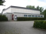 Ein modernes, einstöckiges Verwaltungsgebäude mit weißer Fassade, das von einem großen gepflasterten Vorplatz und einer niedrigen Hecke umgeben ist.