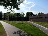 Ein modernes, dunkles Gebäude ist von einem gepflegten Parkbereich mit Wegen und viel Grün umgeben, aufgenommen bei blauem Himmel.