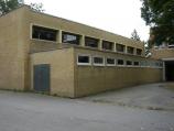 Ein eingeschossiges, hellgelbes Backsteingebäude mit mehreren horizontalen Fenstern, aufgenommen von einem gepflasterten Vorplatz.