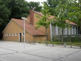 Ein niedriges, eingeschossiges Gebäude mit roten Dachziegeln, angrenzend an eine Fensterfront und umgeben von Bäumen und einem gepflasterten Platz.