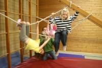 Mehrere Kinder spielen in einer Halle mit einem Seil-Parcours und Hindernisstrukturen.