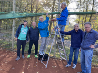 Eine Gruppe von sechs Männern posiert im Freien vor einem Zaun und lehnt sich an eine grüne Stange.