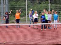 Eine Gruppe von Kindern steht hinter einem Netz auf einem roten Sportplatz.