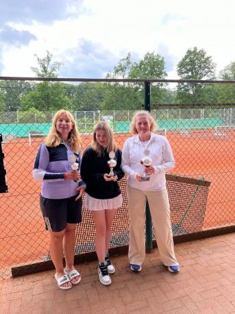 Drei Frauen posieren mit Trophäen vor einem Tennisplatz.