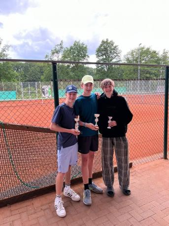 Drei junge Männer stehen vor einem Sportplatz und posieren mit kleinen Trophäen.