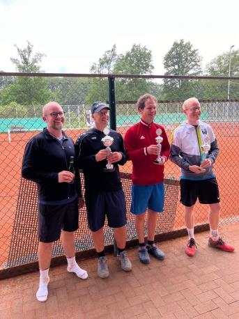 Vier Männer posieren im Freien nach einem Sportevent, wobei drei davon je einen Pokal halten.