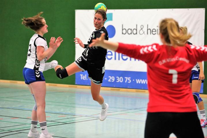 Eine Szene aus einem Handballspiel, bei der eine Spielerin springt, um den Ball zu werfen, während andere Spielerinnen sie umgeben.