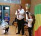 Ein Mann hält einen großen goldenen Pokal und steht inmitten mehrerer Kinder bei einer feierlichen Übergabe in einer Halle.