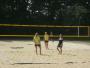 Vier Personen stehen auf einem Sandplatz vor einem Volleyballnetz, hinter dem dichtes Laubwerk sichtbar ist.