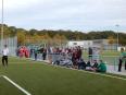 Eine weite Aufnahme eines Kunstrasen-Fußballplatzes mit einer Gruppe von Menschen, die am Spielfeldrand zusammenstehen.