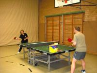 Zwei Männer spielen Tischtennis in einer Sporthalle mit Reihen von Holzschränken an der Wand.