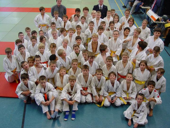 Eine große Gruppe von Kindern, die in hellen Uniformen auf einem Sporthallenboden für ein Gruppenfoto posieren.