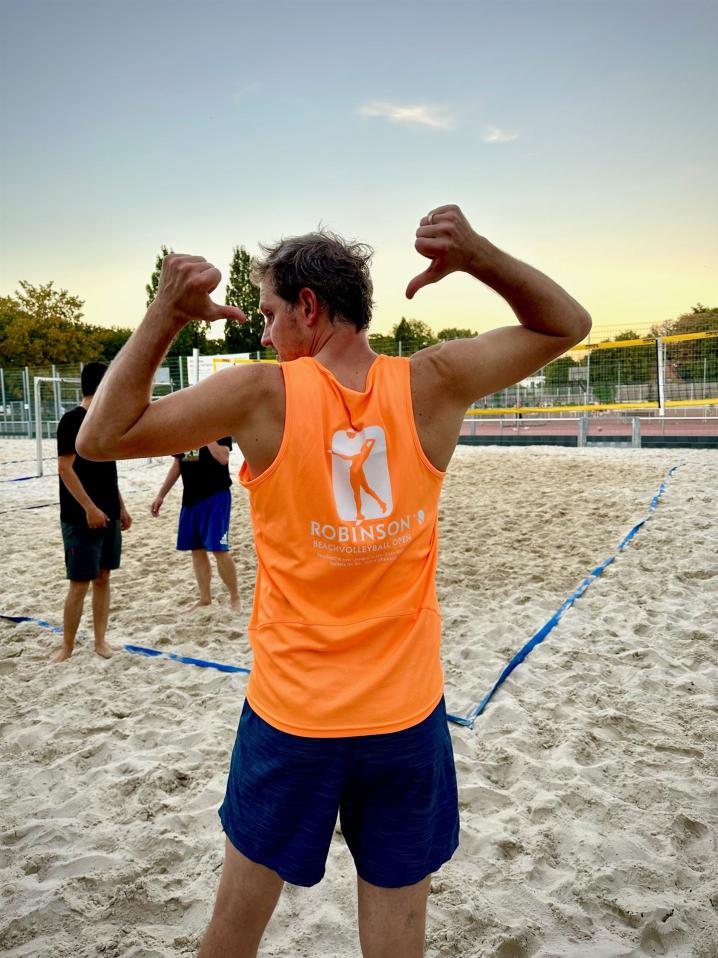 Ein Mann in Sportkleidung posiert mit gestenreichen Händen auf einem Sandstrand.