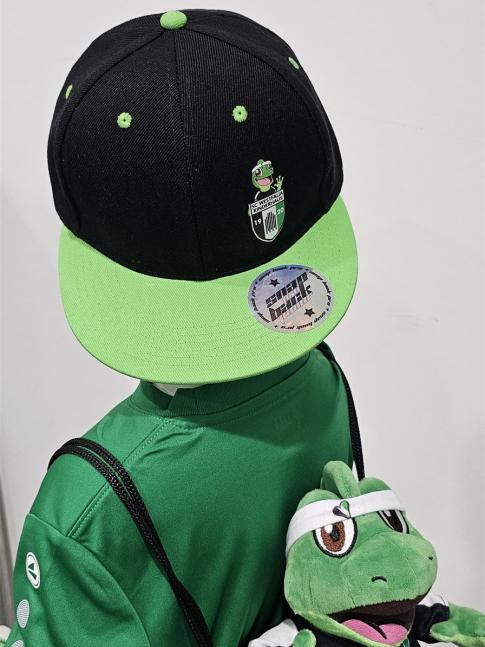 Ein Mann in grünem Shirt und Kappe posiert mit einem Plüschtier eines Froschcharakters.