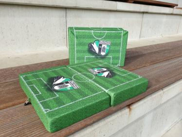 Mehrere grüne Kunstrasen-Matten, die einen Miniaturfußballplatz darstellen, liegen auf einer Holzbank.