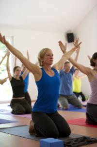 Eine Frau führt in einem Yoga-Kurs eine Dehnübung mit erhobenen Armen aus, während andere Personen in einer Gruppe ebenfalls trainieren.