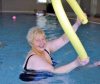 Eine blonde Frau steht lächelnd in einem Schwimmbecken und hält zwei gelbe Schwimmhilfen.