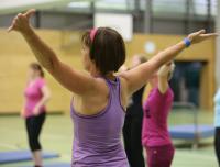 Eine Frau dehnt ihre Arme in einem Fitnessstudio, während andere Personen im Hintergrund trainieren.