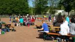 Eine große Gruppe von Menschen ist in einem Park im Freien versammelt, umgeben von vielen Bäumen und Sportanlagen.