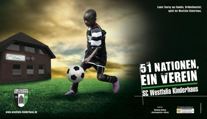 Ein junger Spieler in einem Fußballtrikot steht auf einem grünen Rasen, begleitet von einem Werbeplakat für einen Fußballverein.