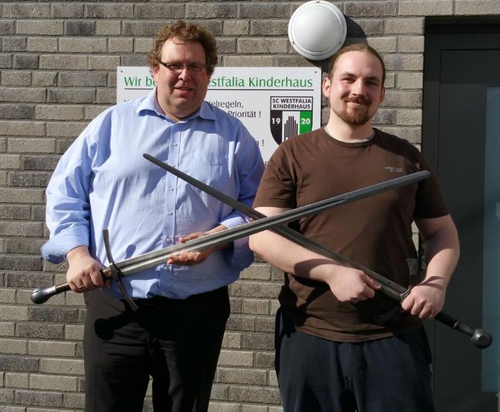 Zwei Männer vor einer Mauer, die jeweils ein langes, zweischneidiges Schwert halten.
