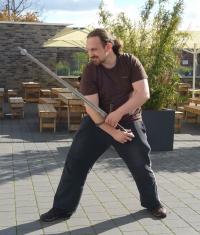 Ein Mann in einer Kampfhaltung mit einem langen Stab oder Speer im Freien.