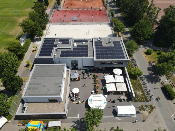 Luftaufnahme eines großen Sport- und Veranstaltungskomplexes mit Solarpaneelen und einem angrenzenden Sportplatz.